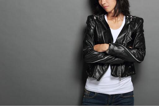 A woman wears a black leather jacket and is leaning against a gray-colored wall.